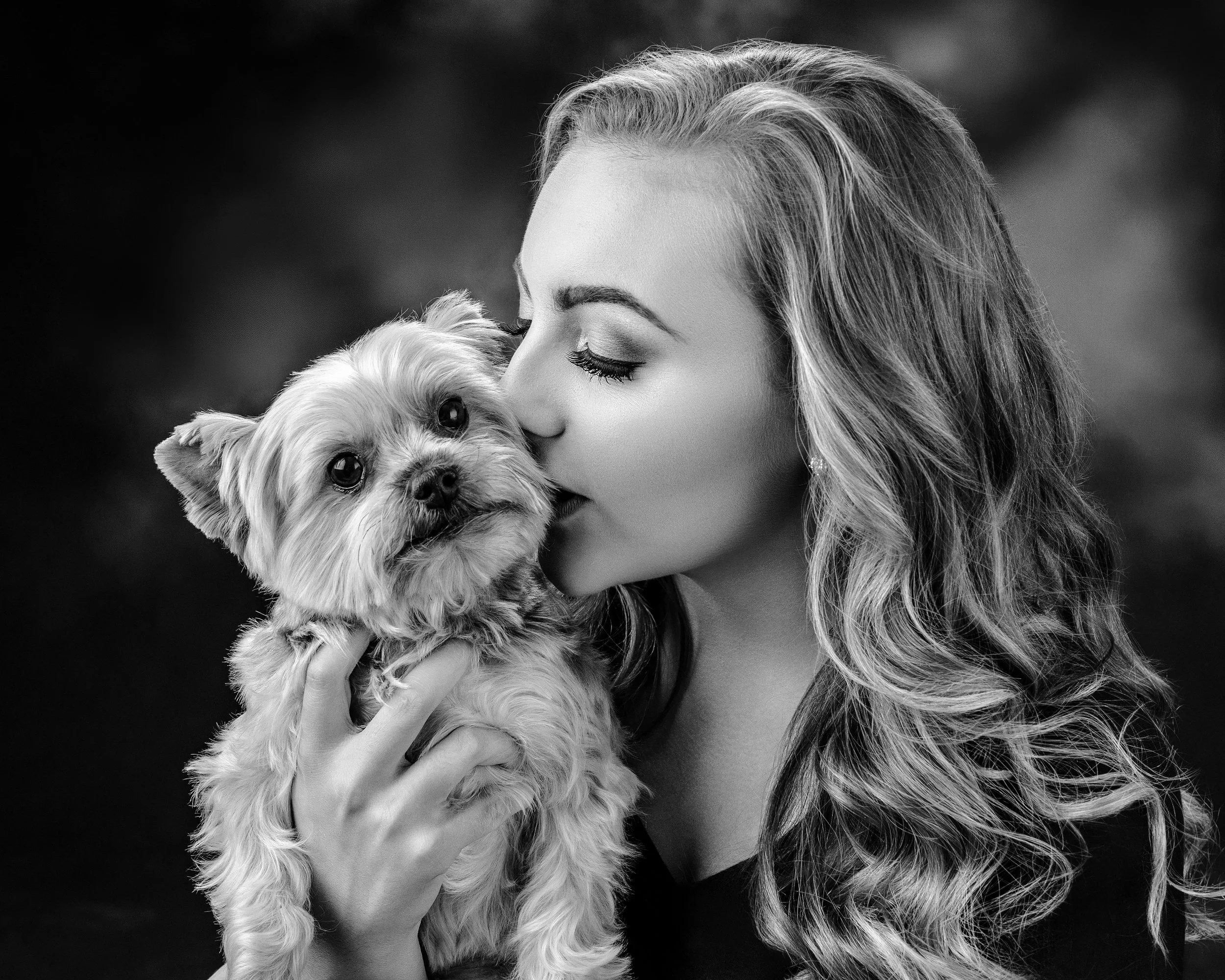 A woman with long, wavy hair holding and gently kissing a small, fluffy dog with light-colored fur.