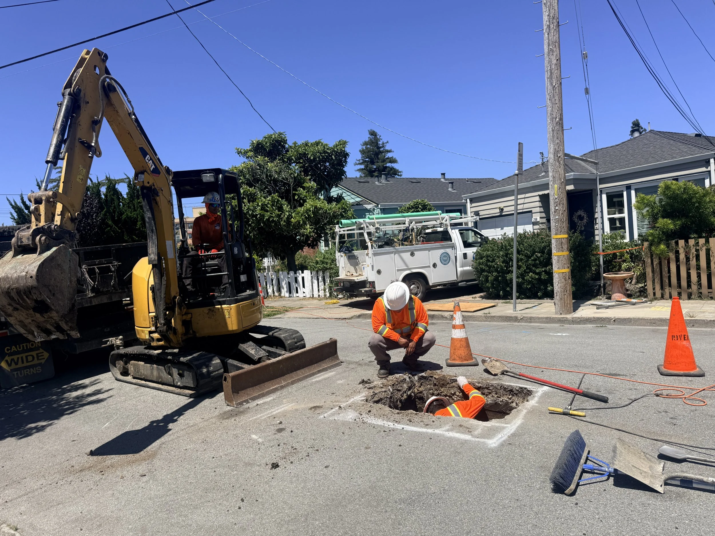 Construction workers repairing a road with a large hole in the asphalt, surrounded by orange traffic cones, a yellow excavator, and utility poles under a clear blue sky.