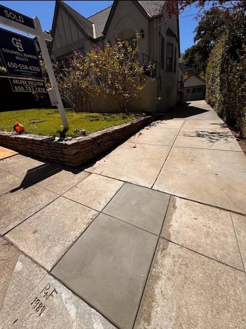 Sidewalk with a newly poured concrete square section, in front of a house with a stone-bordered lawn and a "Sold" sign with realty contact info. There are shadows, bushes, and a driveway leading to a garage in the background.