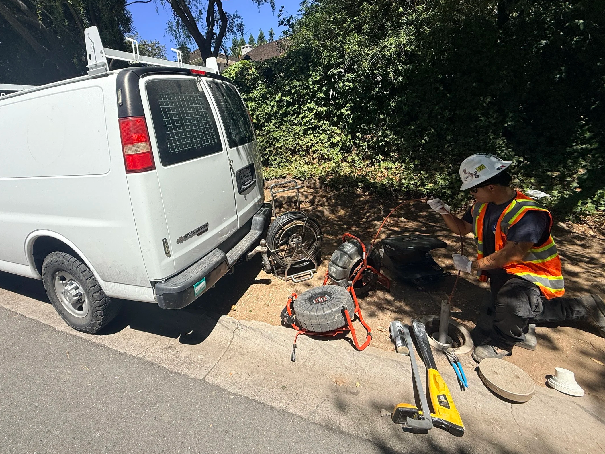 A worker wearing a white safety helmet, sunglasses, and a neon orange safety vest is kneeling on the ground, working on a piece of electrical or machinery equipment. There is a white utility van parked nearby with the rear door open, various tools, a