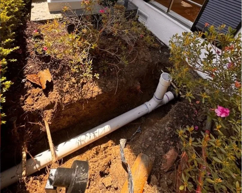 Underground pipe installation next to a house, with soil and plants around.