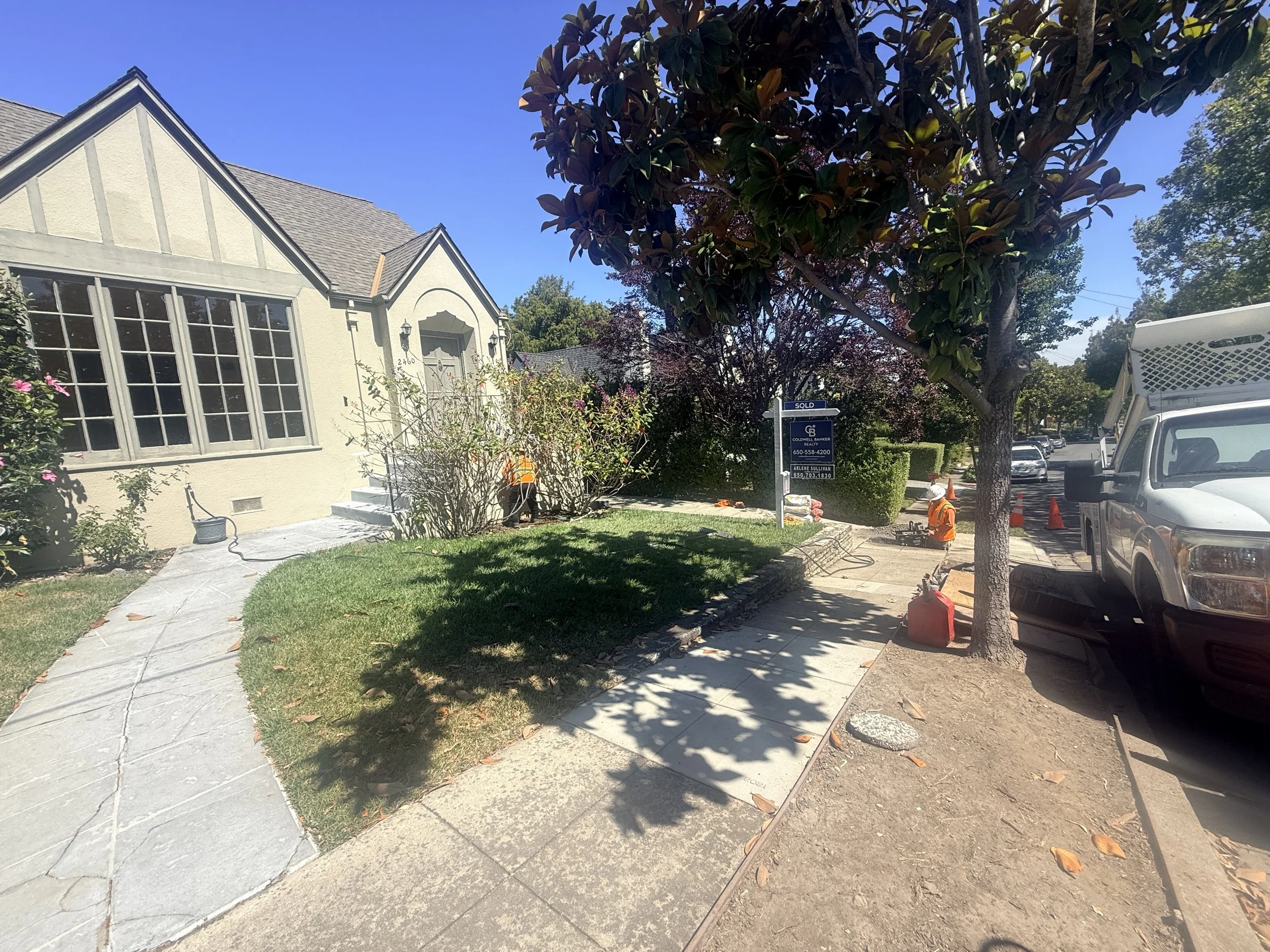 Sidewalk with freshly laid concrete, a house with large windows and a door, trees and bushes, two workers in orange safety vests, and a street with parked cars and orange traffic cones.