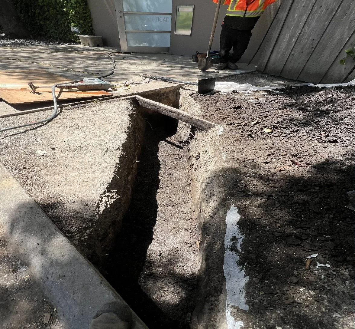 Construction site showing a large trench dug into the ground with a worker wearing a safety vest and helmet, using a tool near a boarded-up doorway and construction equipment around.