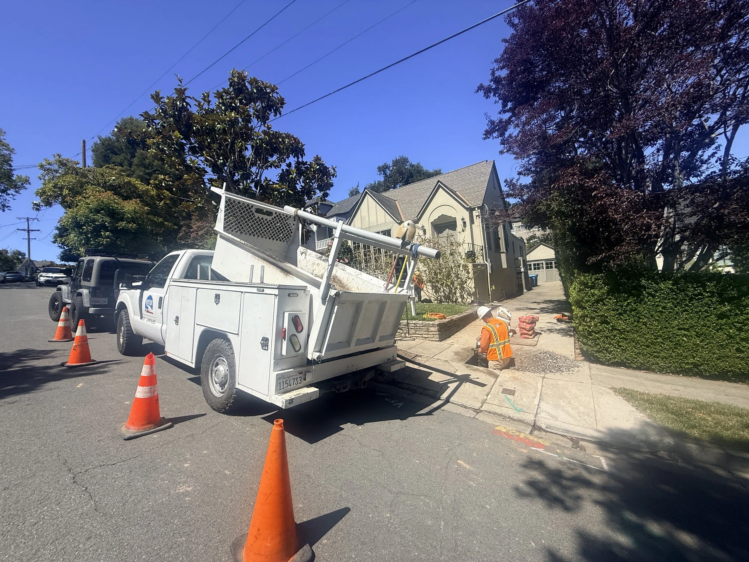 Construction workers are working on a sidewalk, with orange traffic cones and a utility truck in the street. There are trees, houses, and power lines in the background under a clear blue sky.