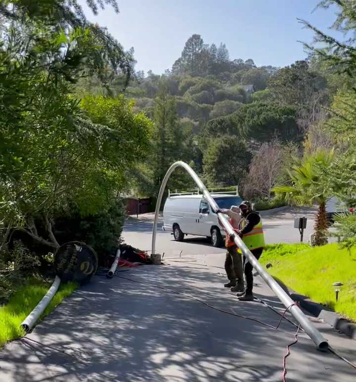 Workers in safety vests repairing a street light pole near a sidewalk with trees and a van in the background.