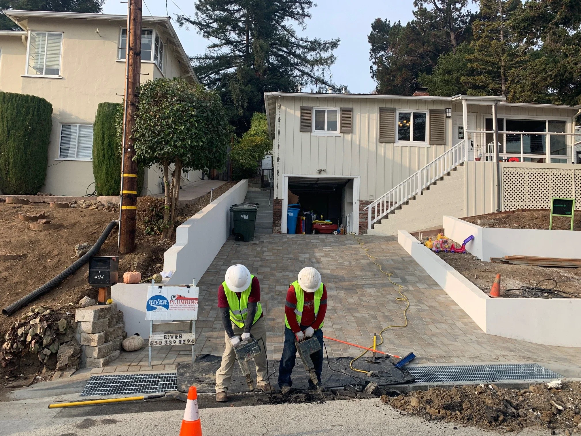 Construction workers paving a driveway in front of a house, with tools, orange traffic cones, and construction signs visible.