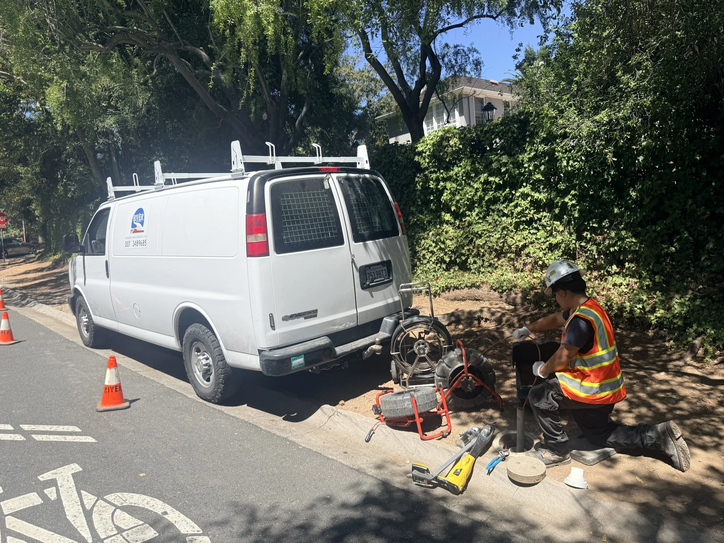 Worker in a safety vest and helmet working on underground utility or pipe line, with a white utility van labeled 'River' parked nearby and traffic cones marking the work area.