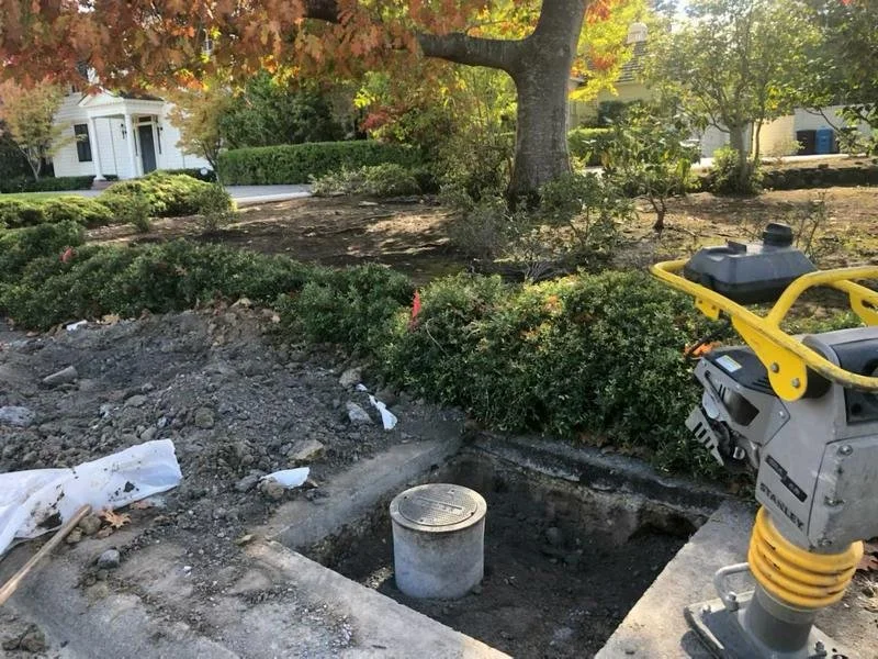 View of a landscaping or construction site in a residential neighborhood with a large tree and bushes, an open hole with a concrete pipe, and construction equipment nearby.