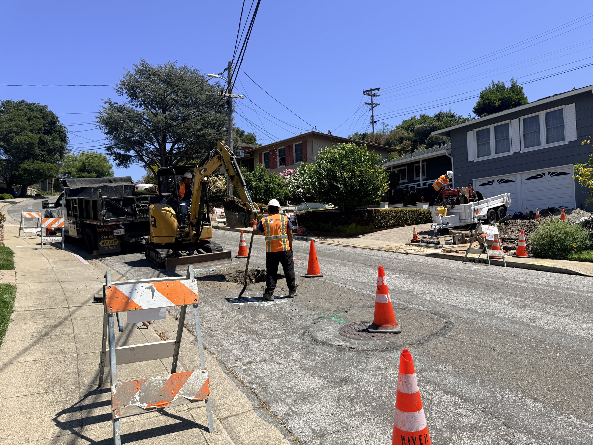 Construction workers and equipment working on a street with orange cones, a small excavator, and a truck in front of residential houses, with trees and power lines overhead under a clear blue sky.