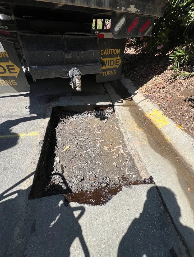 A section of road where asphalt has been removed, revealing a rectangular hole with exposed dirt and some water underneath, next to a large truck with caution signs.