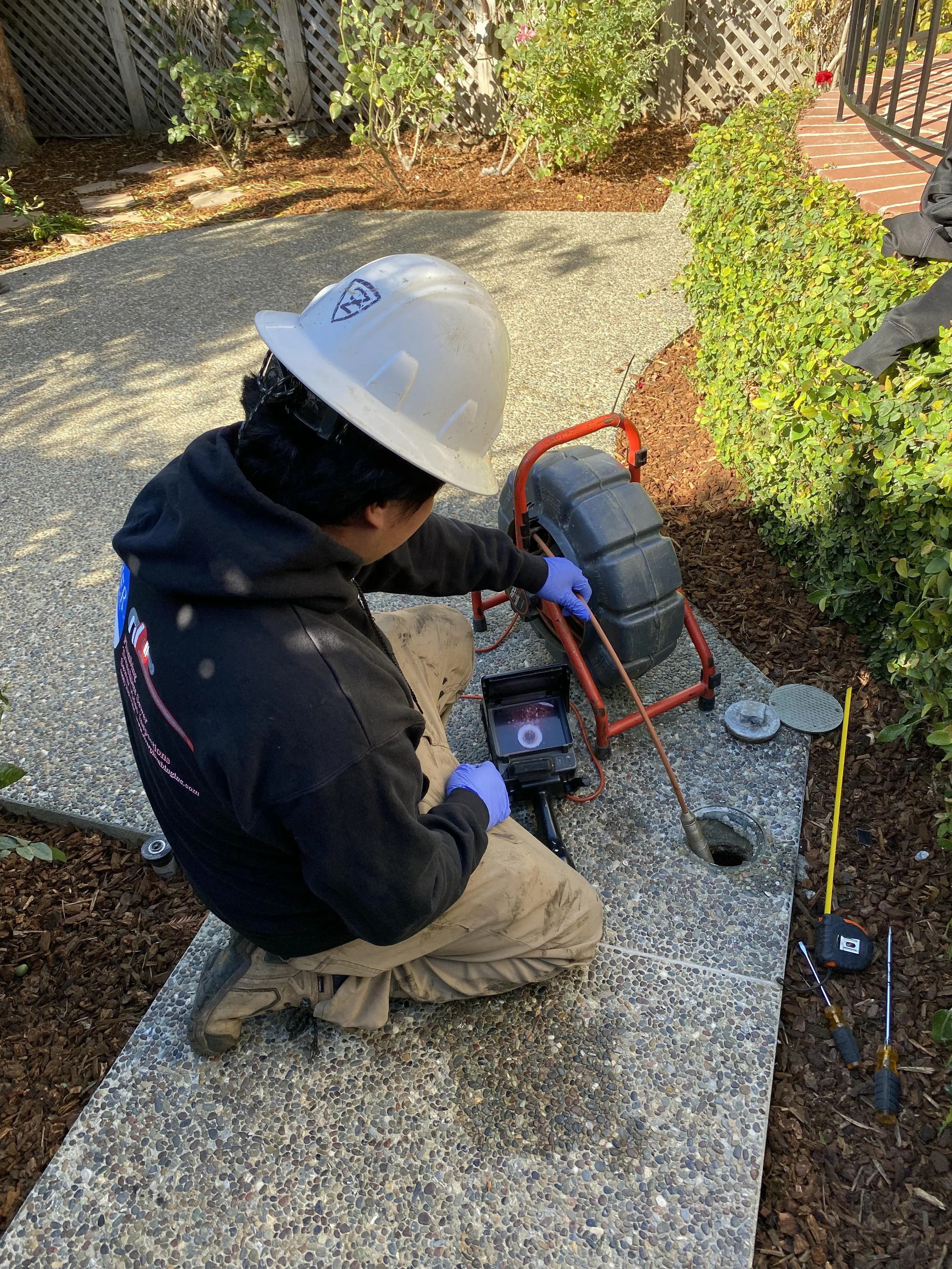 A worker wearing a white safety helmet, black jacket, and gloves is kneeling on a stone surface outside, working with specialized equipment near a hole in the ground. There are various tools and a small screen on the ground nearby.