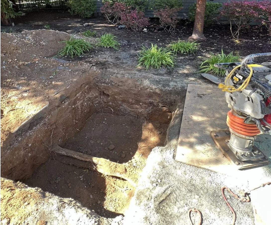 A large rectangular hole in the ground, possibly for installing a post or foundation, with a power tool and construction equipment nearby in a backyard garden area with plants and a tree.