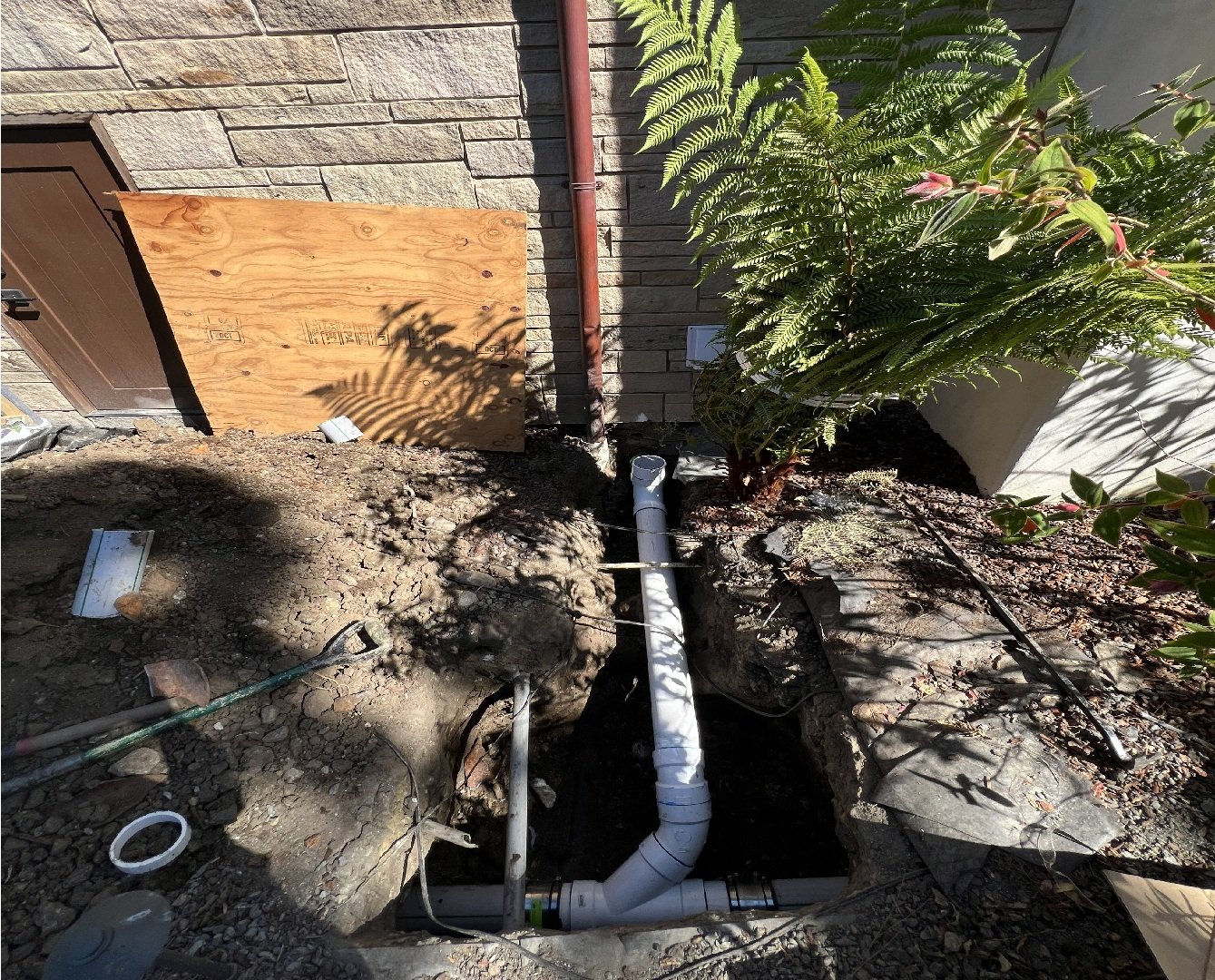 Construction site showing an open trench with PVC piping, a wooden board blocking part of the site, and a brick wall with a door, plants, and shadows.