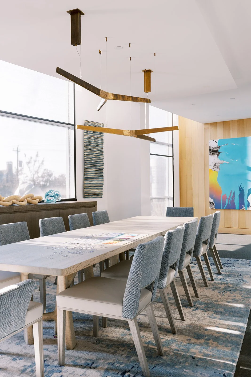 Modern conference room with large wooden table, grey upholstered chairs, abstract colorful artwork, and geometric ceiling light fixture.