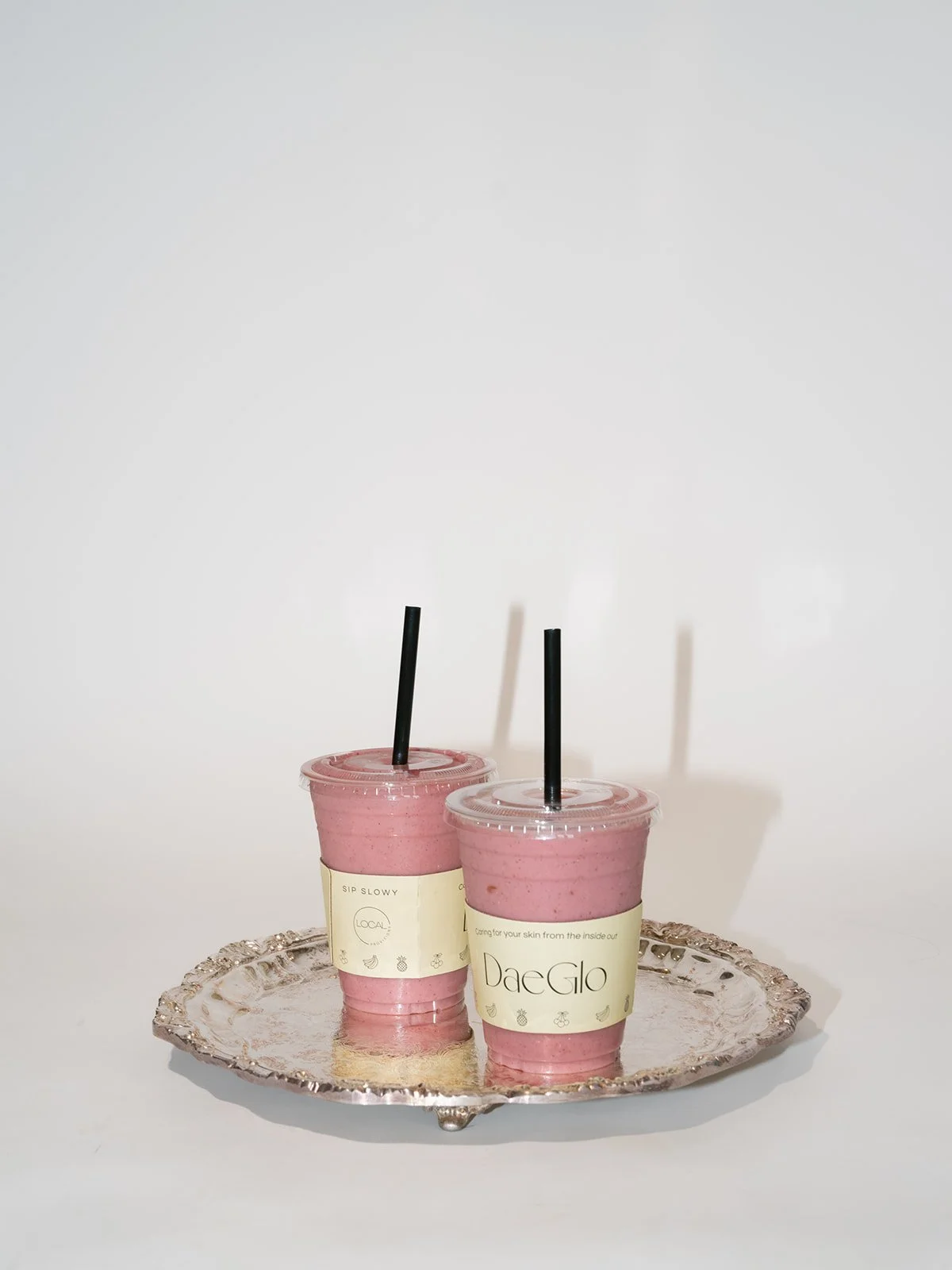 Two pink smoothies in clear plastic cups with black straws placed on a decorative silver tray against a plain white background.