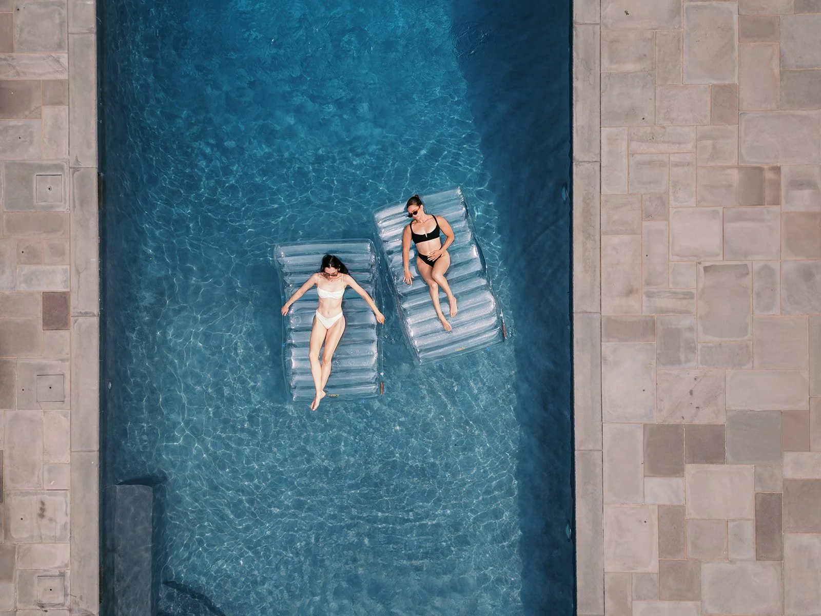 Two women lying on inflatable loungers in a swimming pool, viewed from above. One woman is wearing a black bikini and sunglasses, and the other is in a white bikini. The pool is bordered by stone tiles.