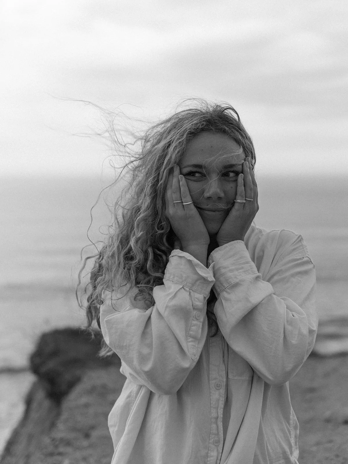 A woman with curly hair in a light-colored shirt standing outdoors near the water, holding her face in her hands and smiling. The background shows a blurred ocean and cloudy sky.