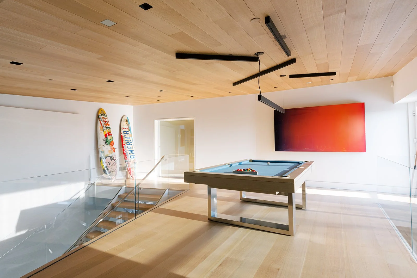 A modern recreational room with a pool table, two skateboards against the white wall, a large flat-screen TV, and a staircase with glass railing. The ceiling is wooden with black light fixtures.