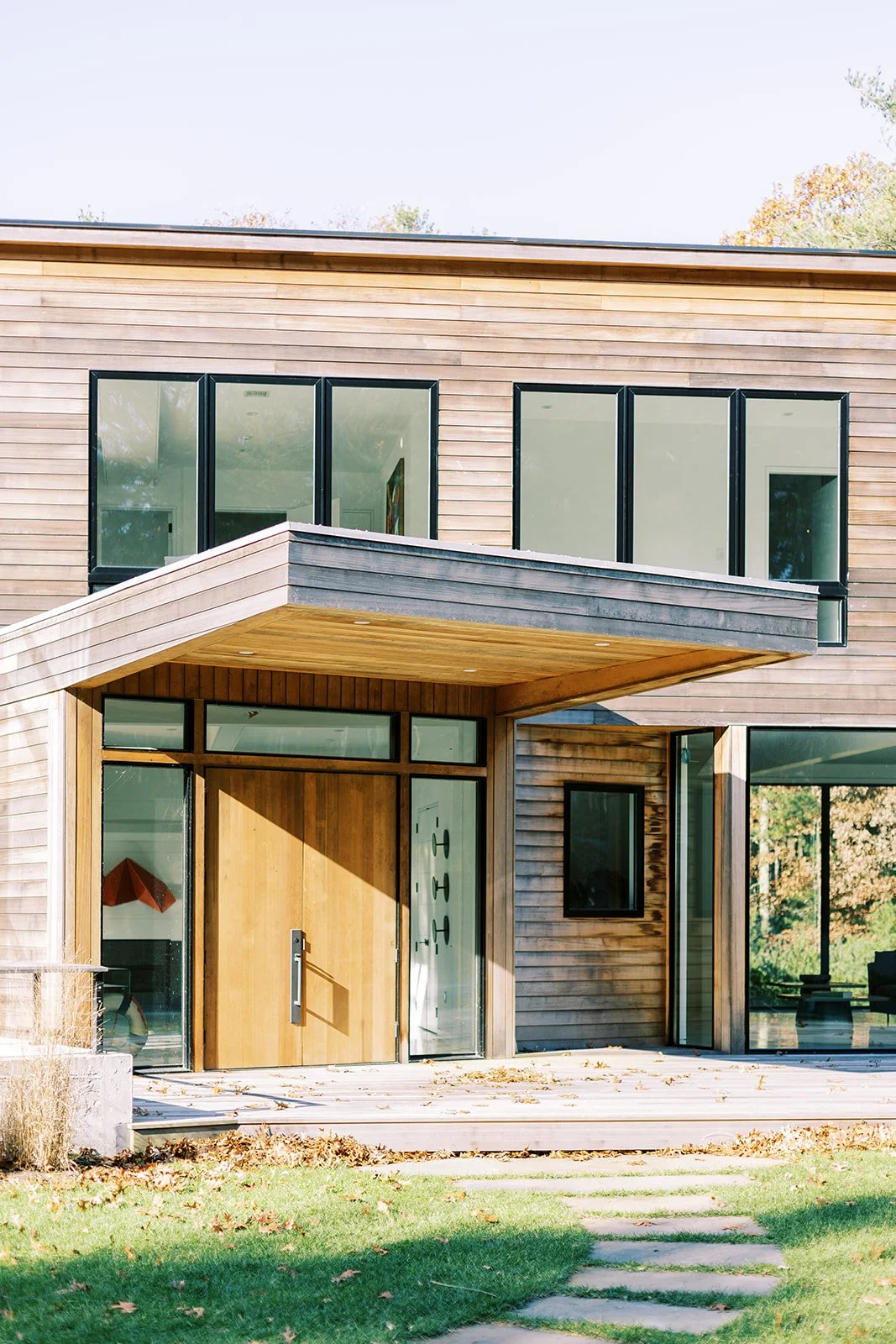 Modern house with wooden exterior, large glass windows, and a wooden front door, surrounded by a lawn and a stone pathway.