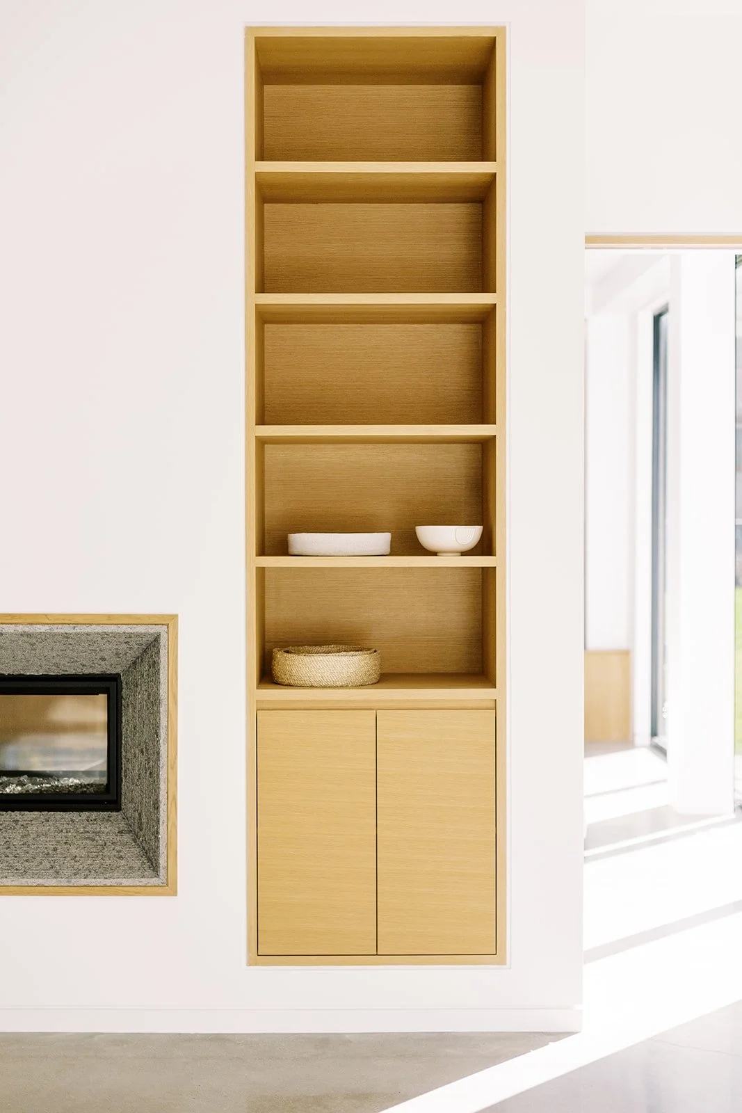 A minimalist wooden shelving unit built into a white wall with three open shelves, two of which contain decorative bowls and a basket, and a closed cabinet at the bottom.
