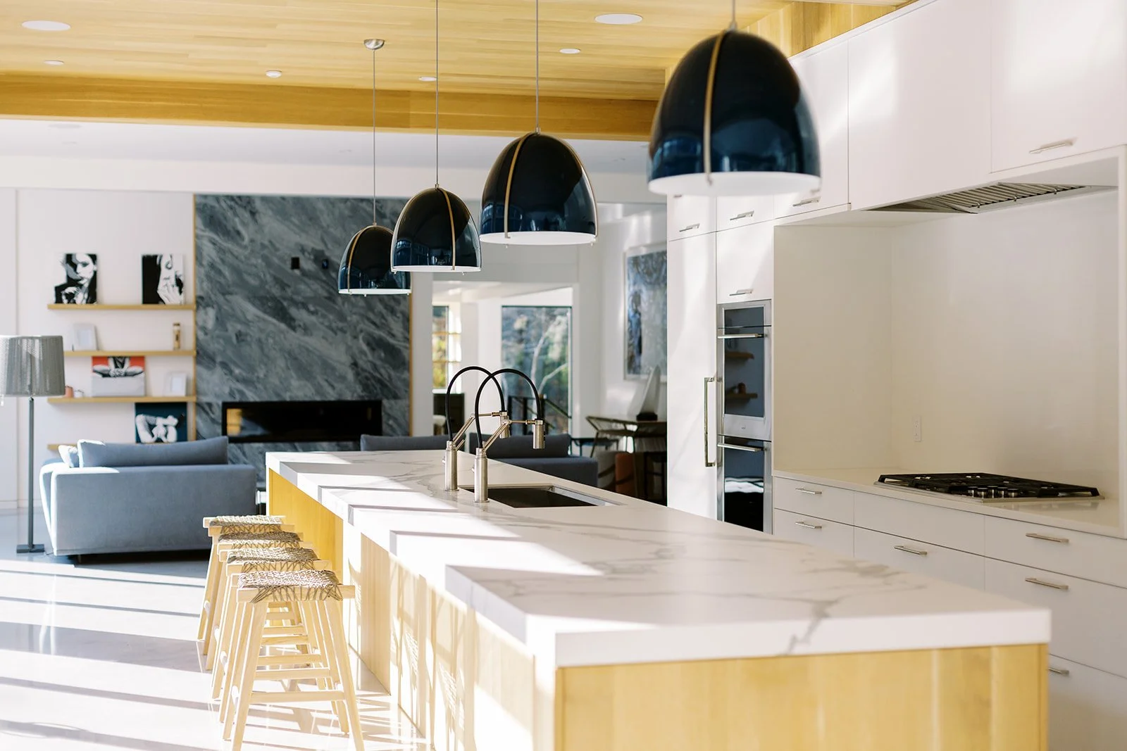 Modern kitchen with white marble island, black pendant lights, and a living room in the background with sofas and artwork.