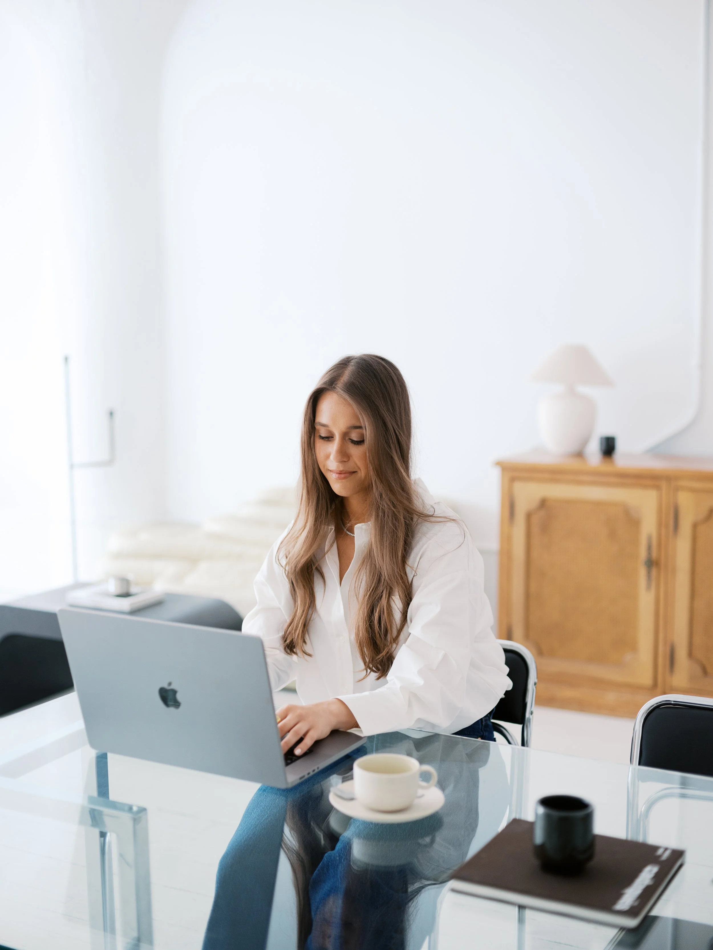 A young woman with long brown hair, sitting at a glass conference table, working on a silver laptop. She is wearing a white shirt. On the table are a coffee cup, a black notebook, and a pen. The room has white walls and a wooden cabinet with a decorative lamp in the background.