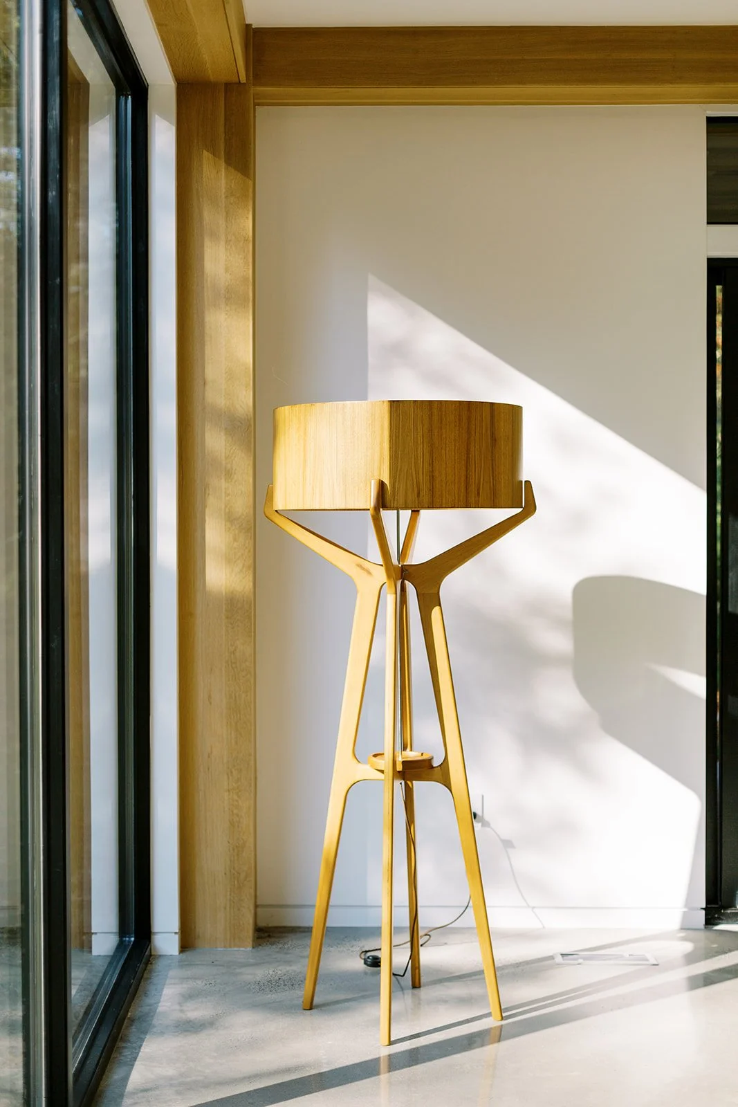 A wooden floor lamp with a lampshade, positioned in a modern interior near a window with sunlight casting shadows.