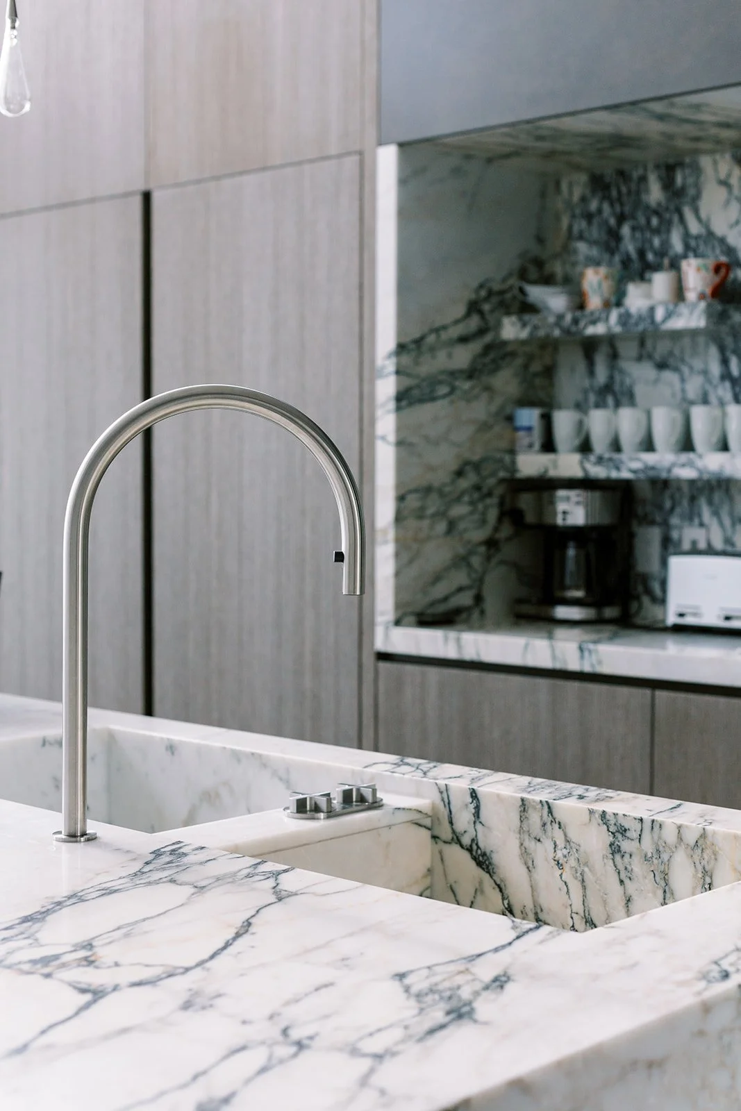 Close-up of a modern kitchen with a marble countertop and backsplash, stainless steel faucet over a sink, and wooden cabinets with a marble shelf holding cups, a coffee maker, and a toaster.