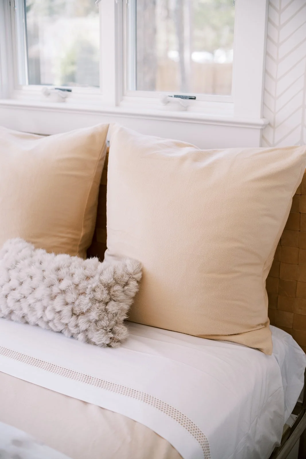 Close-up of a cozy bed with beige pillows, a white furry cushion, and a white blanket in front of a window with sunlight streaming in.