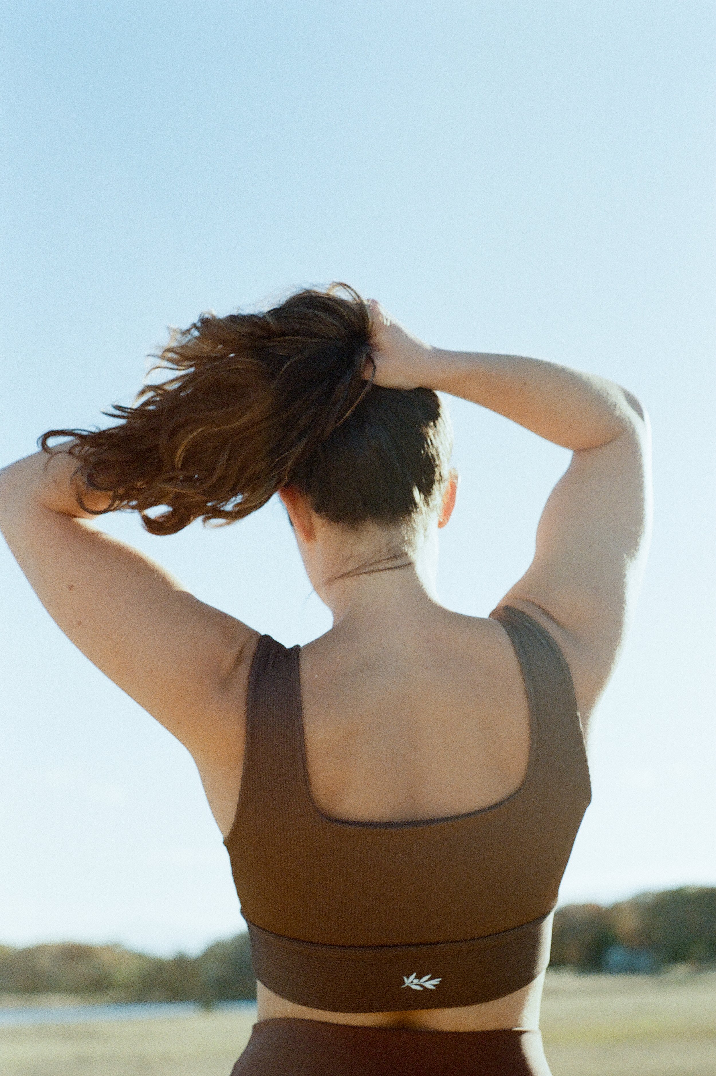 A woman with short hair in a ponytail, wearing a brown sports bra, stands outdoors with her back to the camera, holding her head with both hands, against a clear sky and distant landscape.