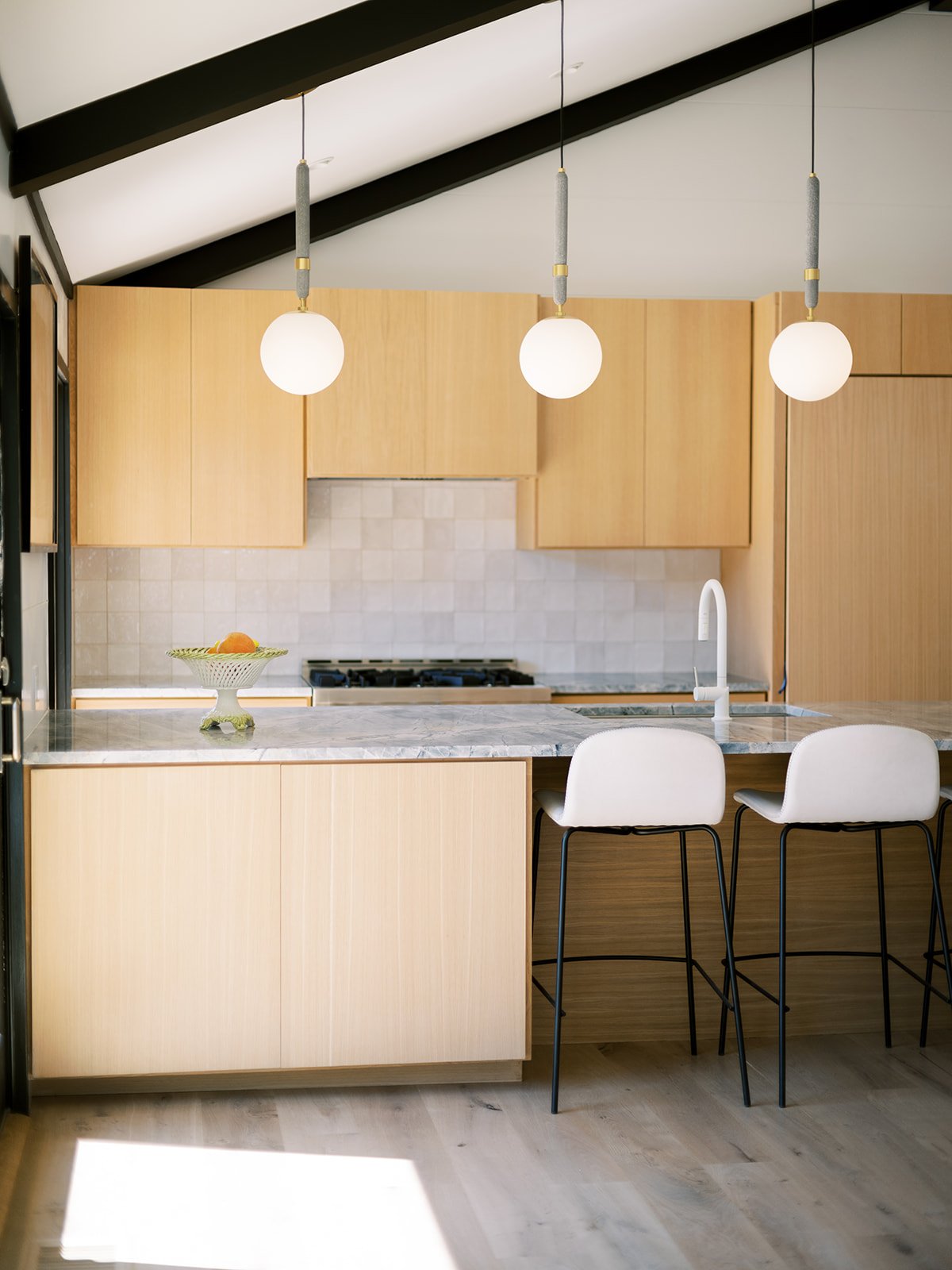 Modern kitchen with wood cabinets, marble countertop, and white stools at a breakfast bar. Pendant lights hanging from a high, sloped ceiling. A bowl with oranges on the counter.