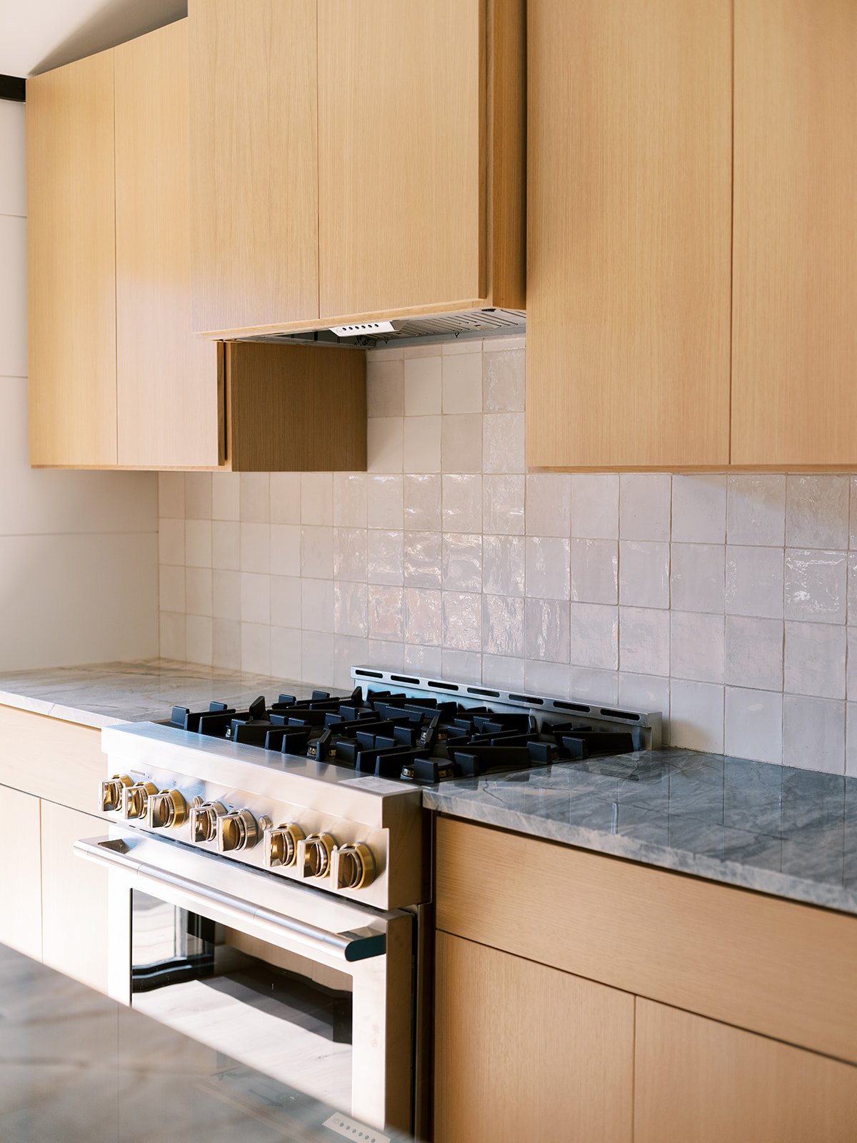 Kitchen with beige cabinets, beige tiled backsplash, marble countertop, and a stainless steel gas stove with gold knobs.
