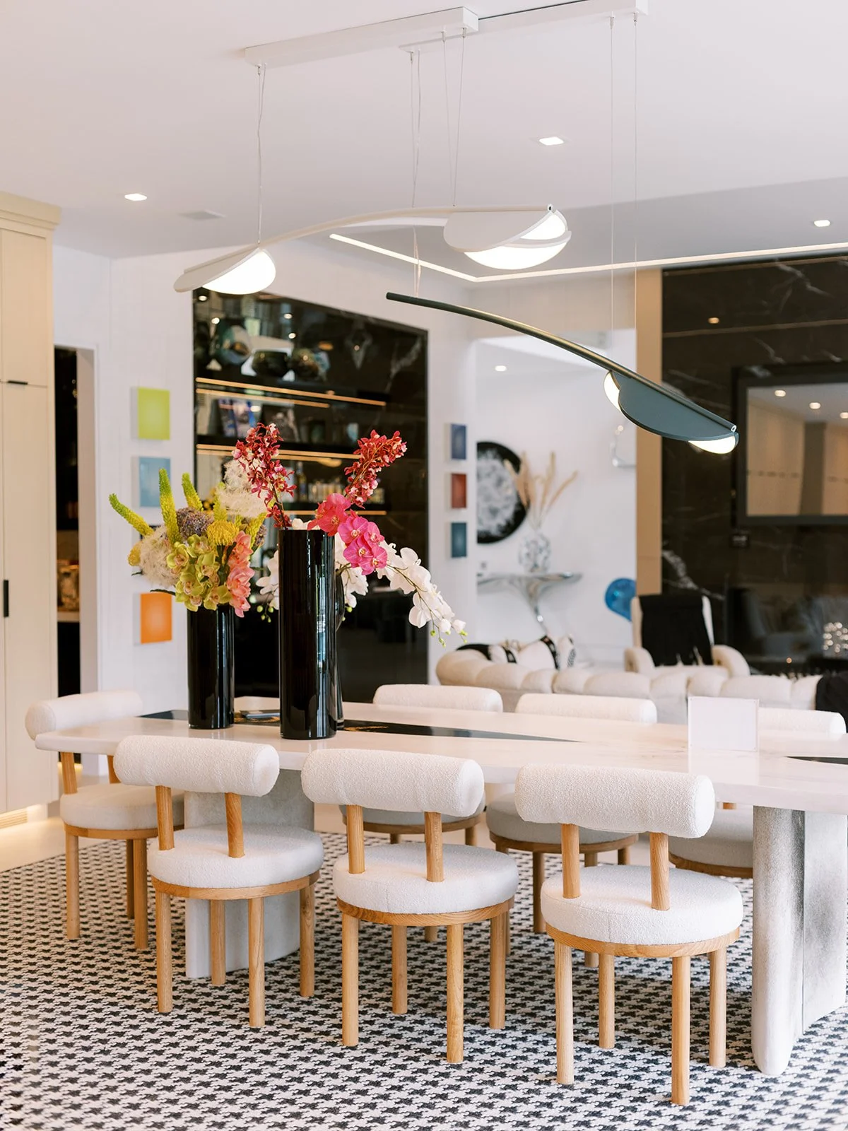 Modern dining room with a white oval table, eight upholstered chairs with wooden legs, and three black vases with pink and green flowers on top. Contemporary lighting fixtures hang above the table with a black and white patterned rug underneath.