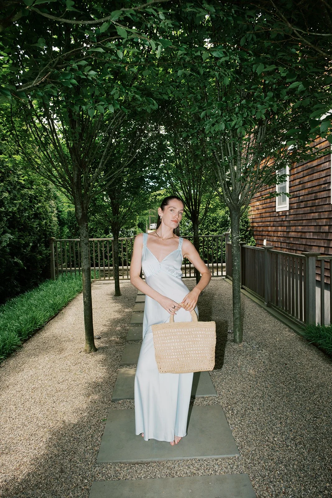 A woman in a white dress holding a beige woven bag standing on stone tiles in a garden with trees and a wooden fence.