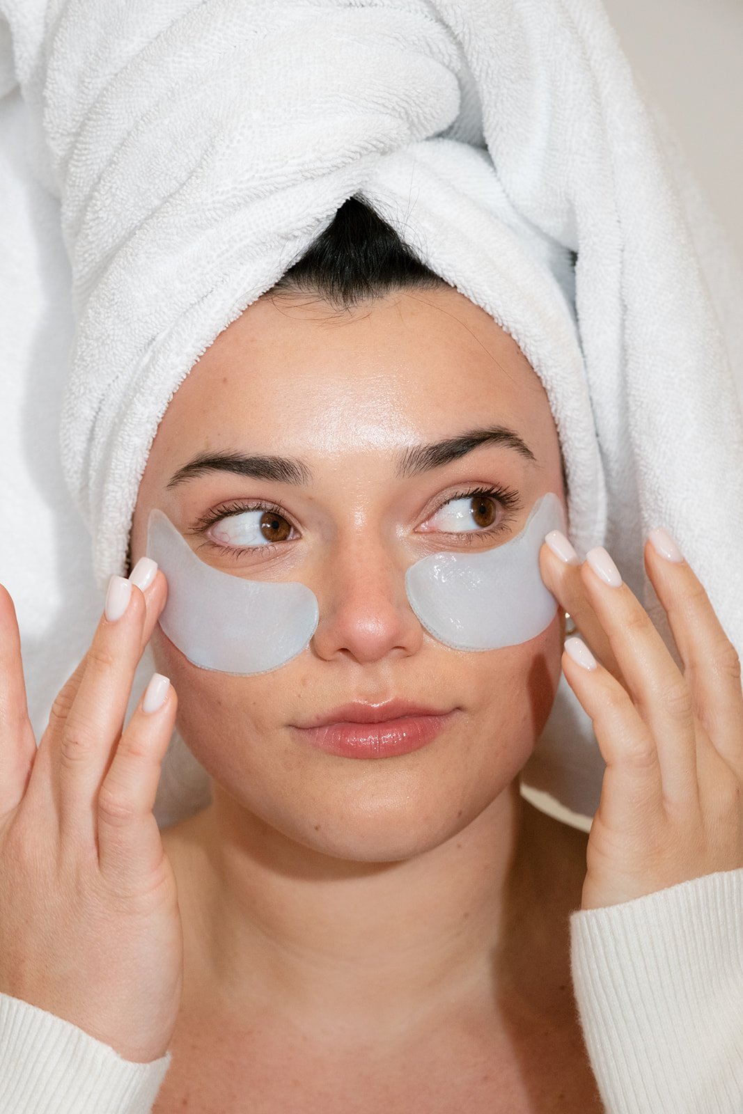 A woman with a towel wrapped around her head applying under-eye patches.