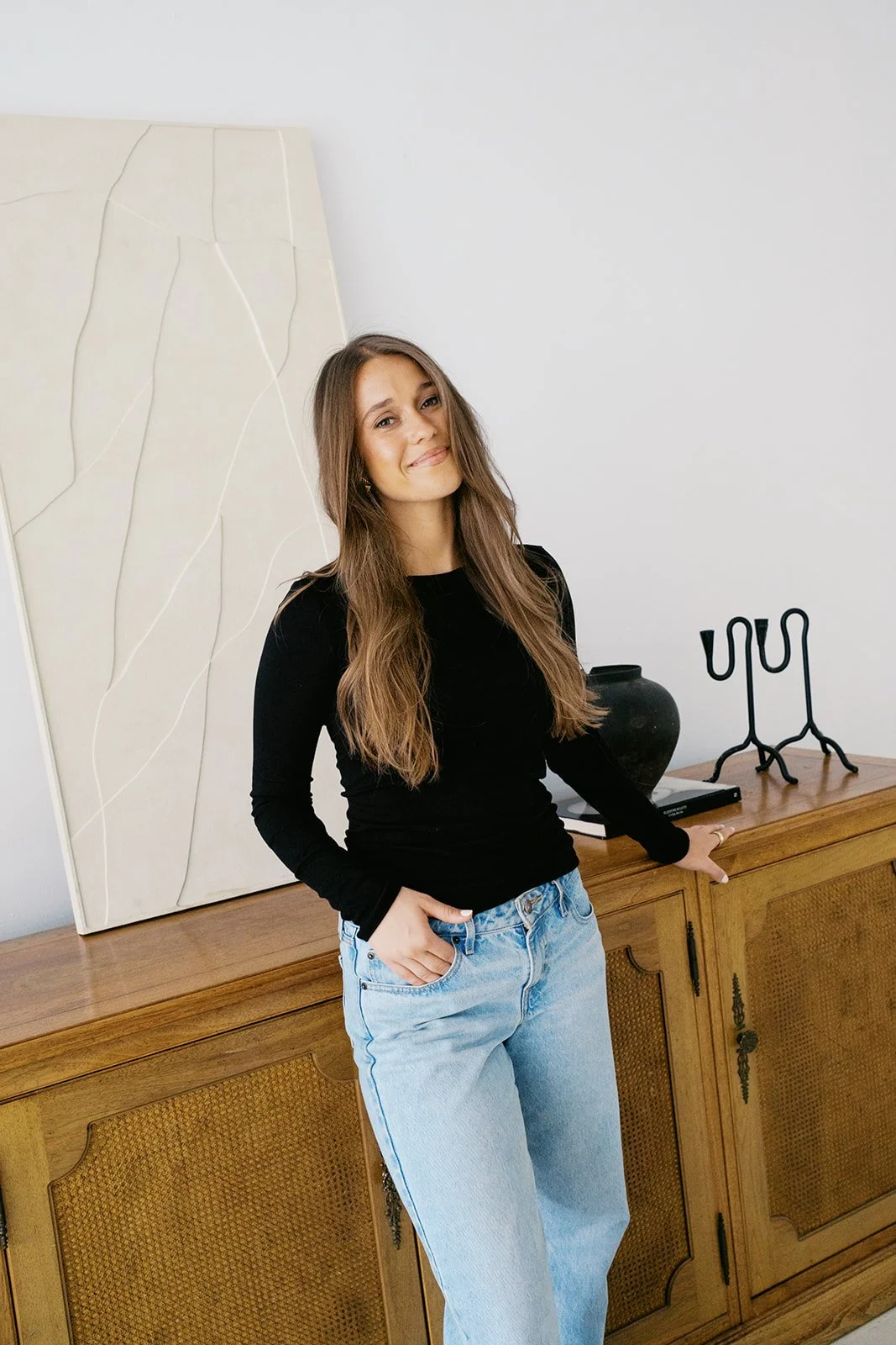 A young woman with long brown hair, wearing a black long-sleeve top and light blue jeans, is standing next to a wooden sideboard with her right hand resting on it, smiling slightly at the camera in a brightly lit room with minimal decor.