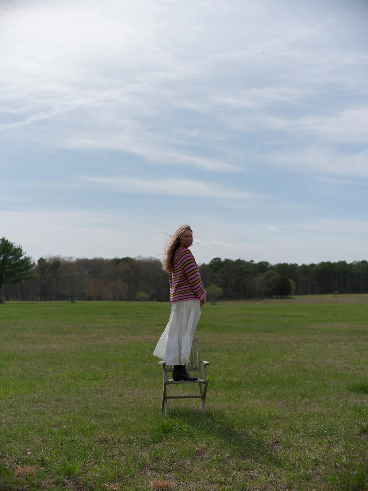 A woman with curly hair standing on a chair outdoors in a grassy field with trees in the background and a cloudy sky.