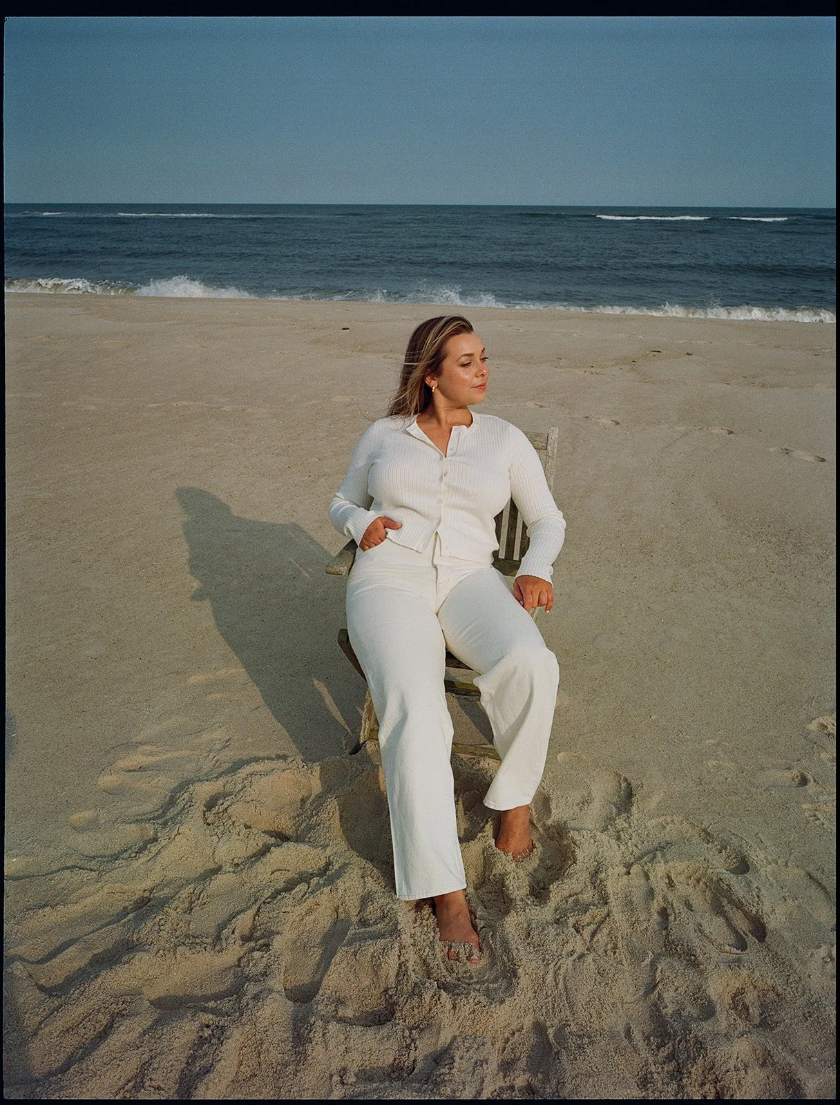 Woman sitting on a beach facing away from the ocean, dressed in white, with her hair blowing in the wind, casting a shadow on the sand.