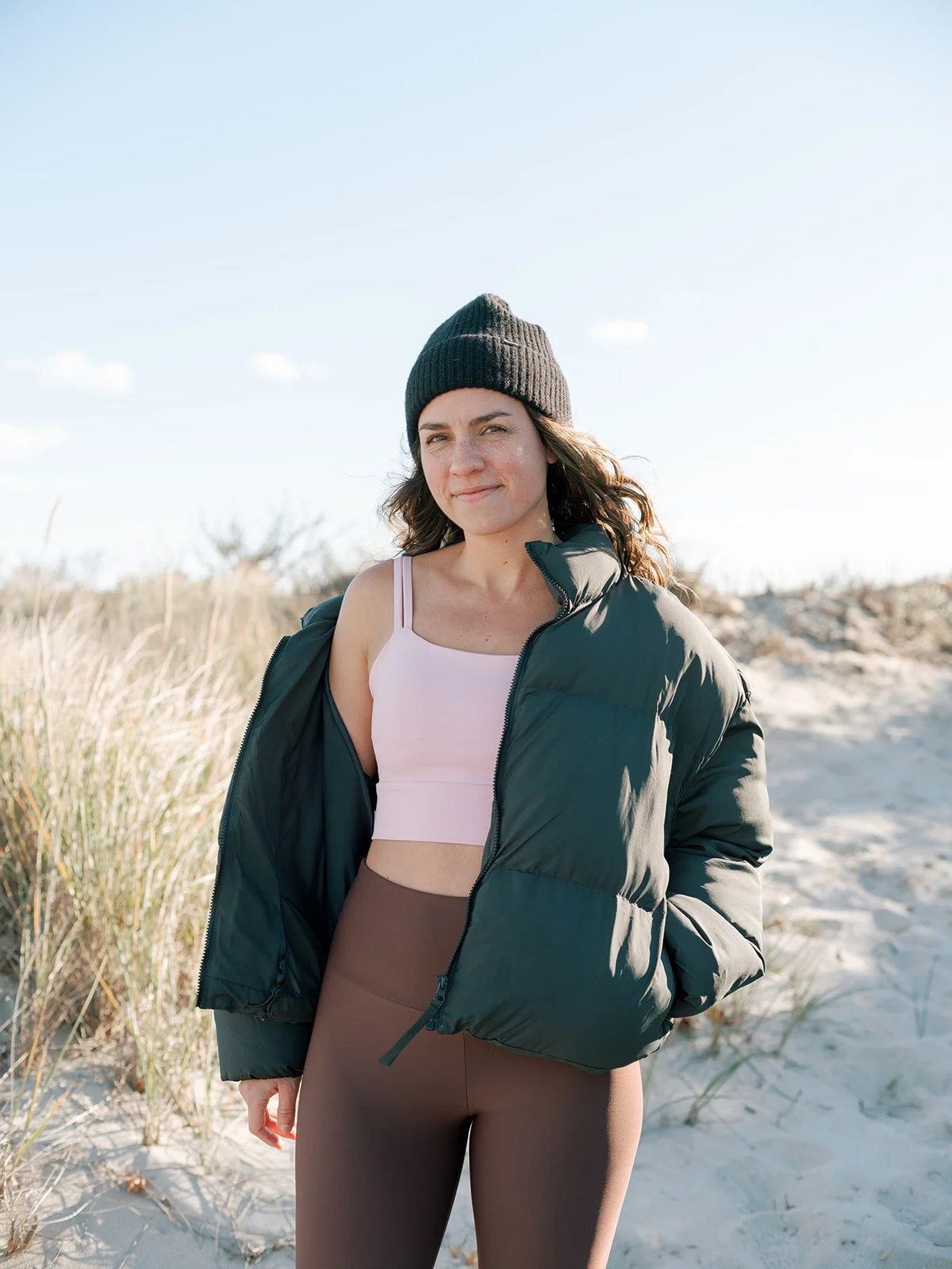 Young woman standing outdoors on a sandy beach with tall grasses, wearing a black beanie, light pink sports bra, green puffy jacket, and brown leggings, smiling at the camera.