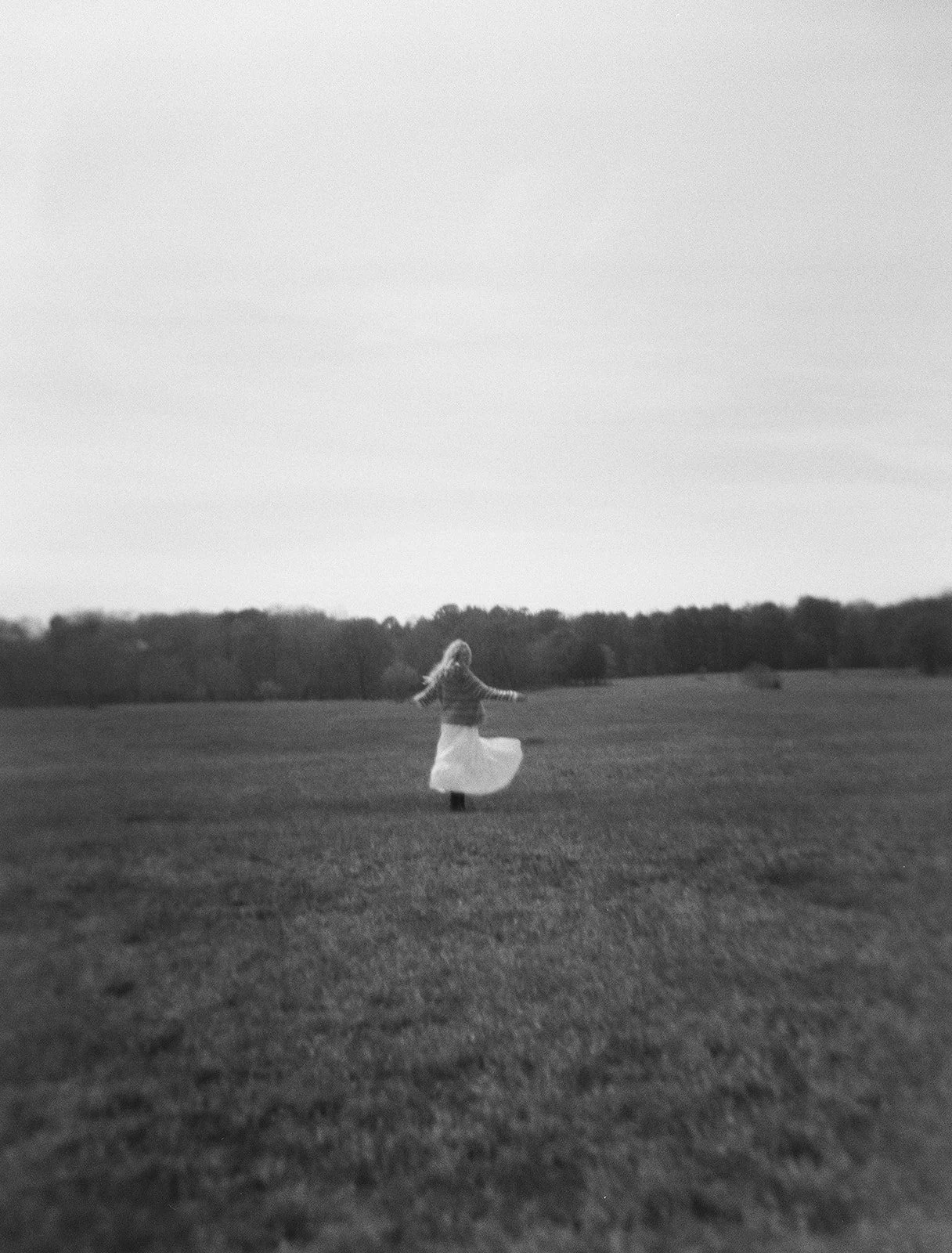 A black and white photo of a woman spinning in a grassy field with her long hair flowing and a line of trees in the background.