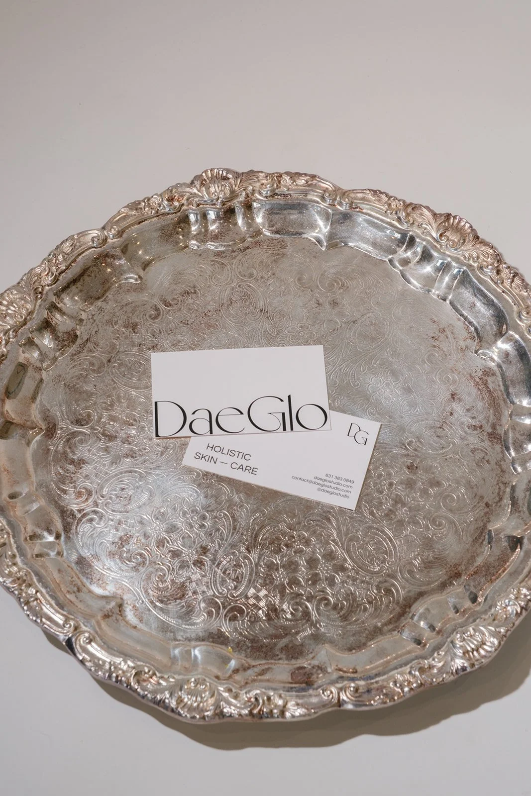 Decorative silver tray with ornate edges and intricate engraved patterns, labeled with a business card for Daeglo Holistic Skin Care.