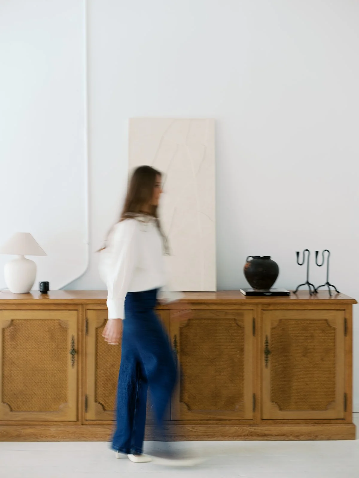 A woman with long brown hair walking past a wooden cabinet with decorative items, including a white lamp, black vase, and metal candle holders, against a white wall with a blank canvas.