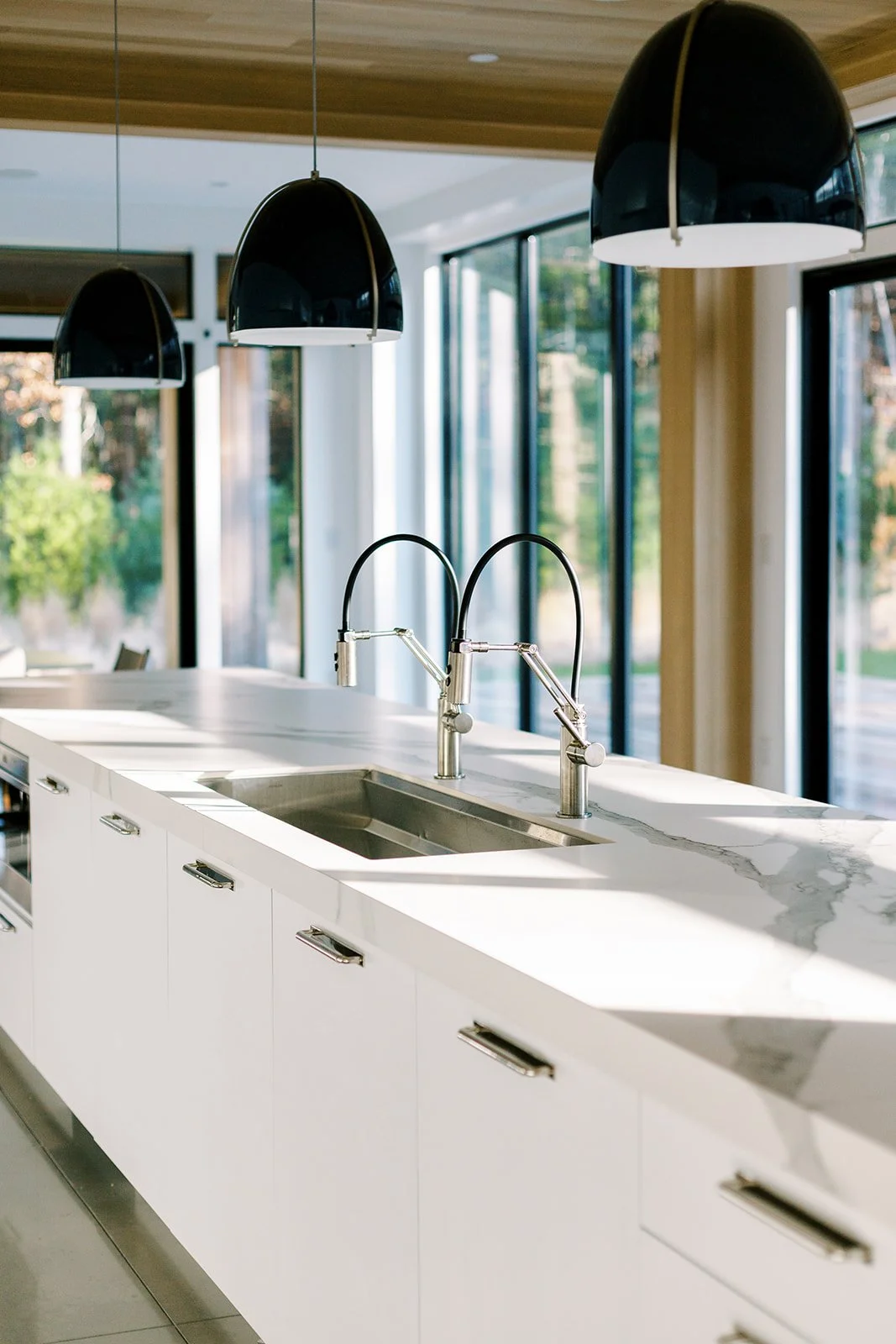 Modern kitchen island with a marble countertop, sink, and dual gooseneck faucets, illuminated by black pendant lights, with large windows and a view of greenery outside.