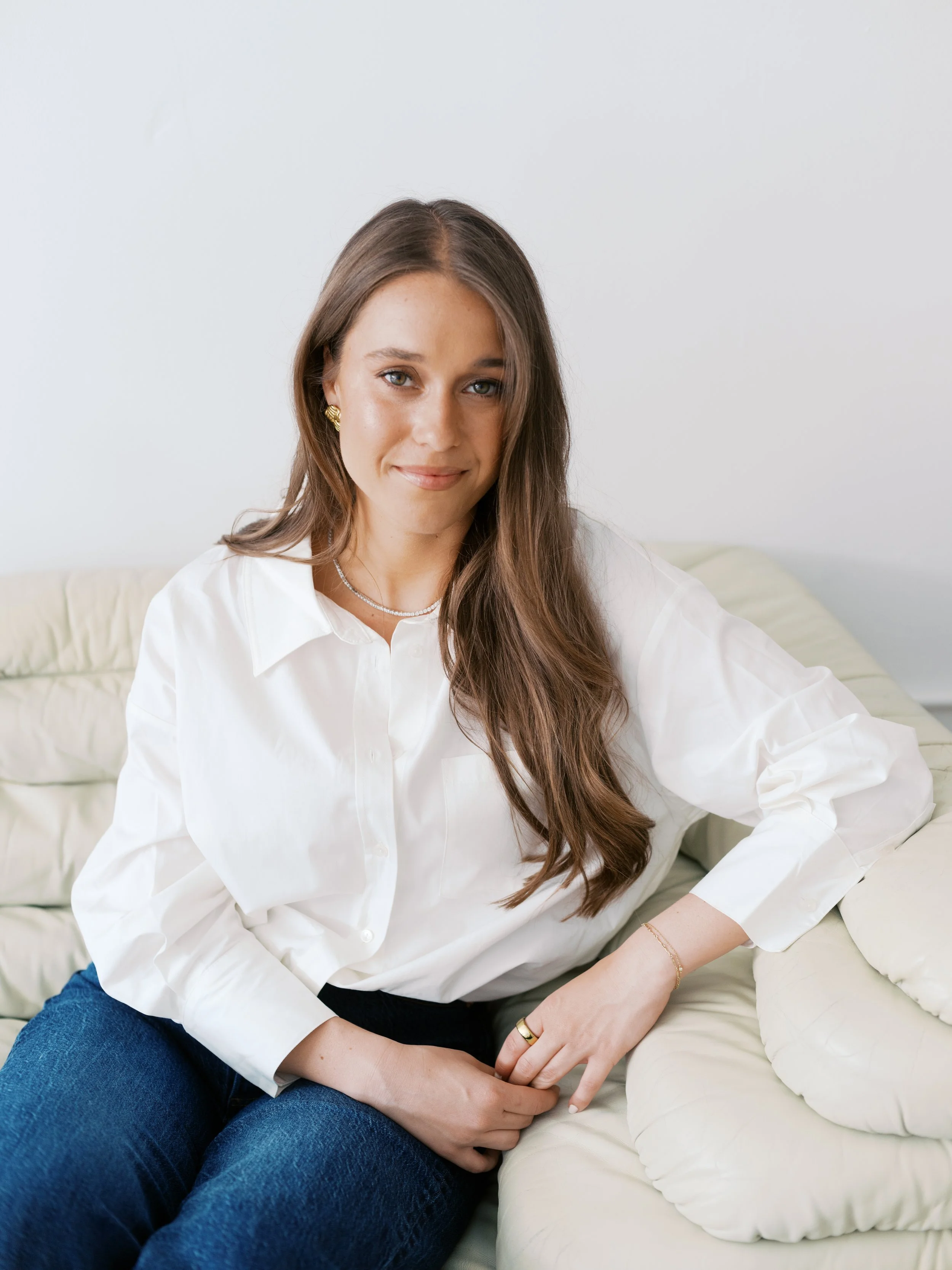 Portrait of a young woman with long brown hair, wearing a white shirt, blue jeans, sitting on a cream-colored sofa, smiling softly with a neutral background.