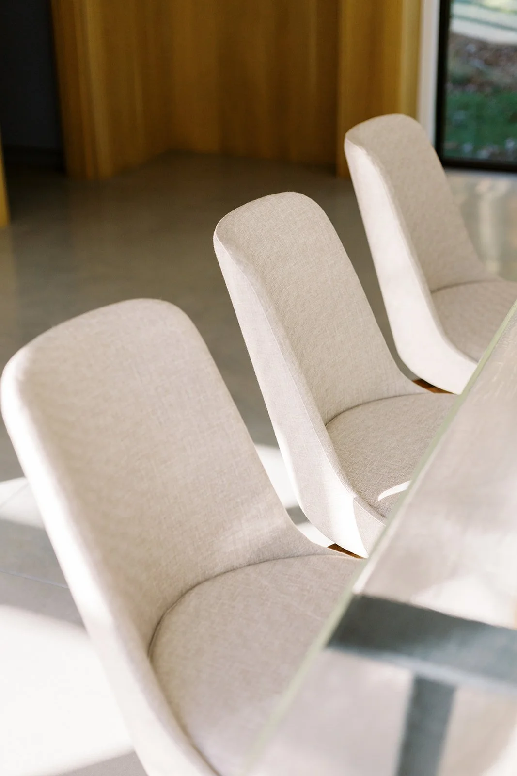 Three beige upholstered chairs in a row at a dining table in a modern room with large windows.