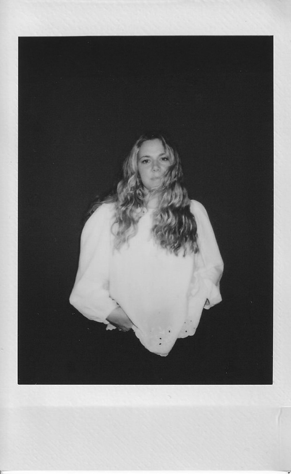 Black-and-white photo of a woman with long, wavy hair, standing against a dark background, wearing a light-colored top with puffed sleeves.