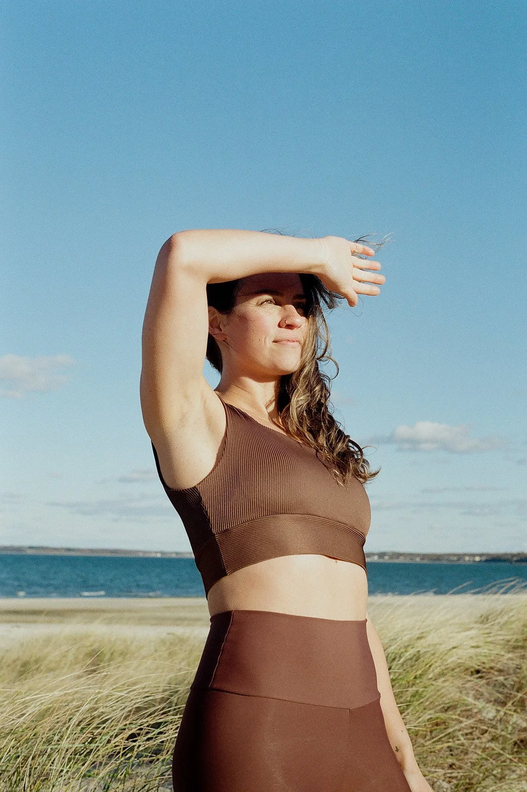 Woman in athletic wear standing outdoors on a beach, shielding her eyes from the sun, with water and sky in the background.