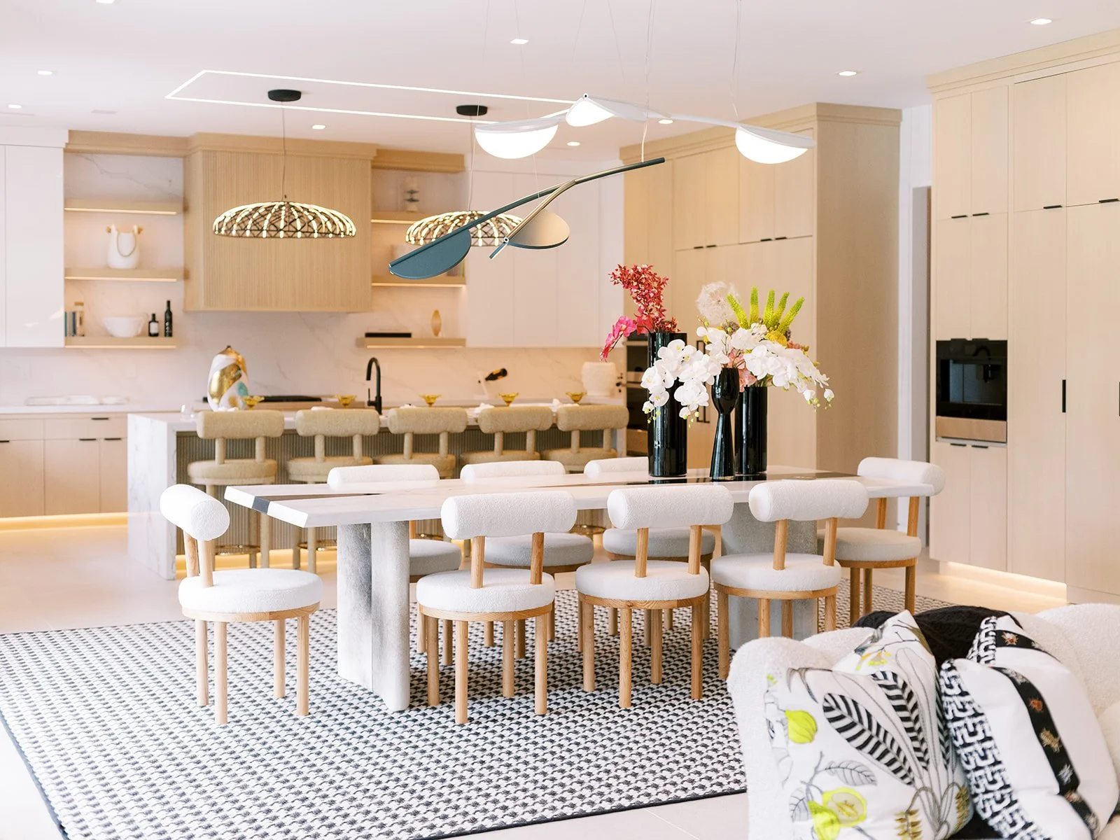 Modern, bright kitchen and dining area with a large white dining table, eight white chairs, a floral centerpiece with pink and white flowers, beige cabinetry, marble backsplash, and unique lighting fixtures.