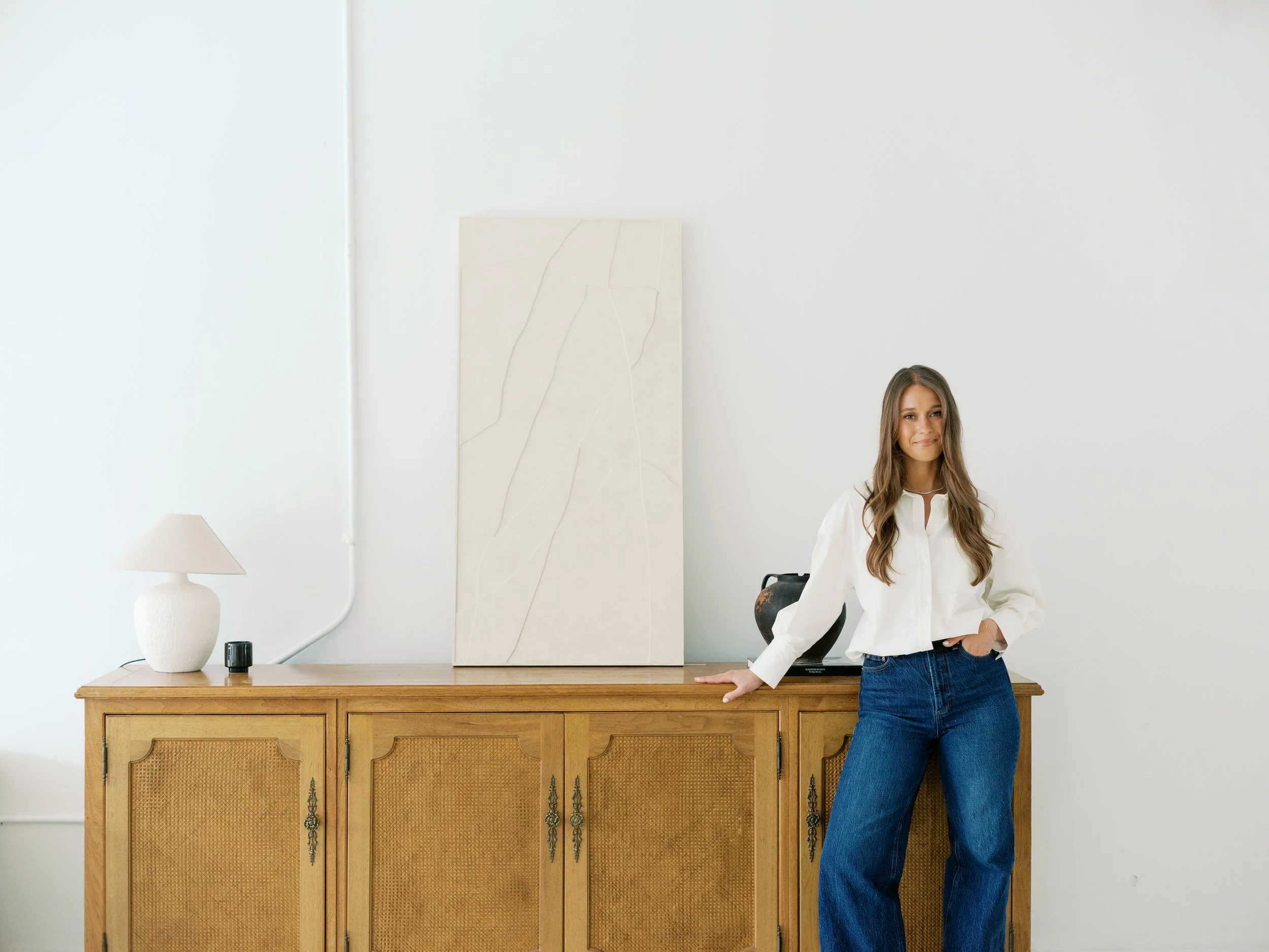 A woman with long wavy brown hair in a white blouse and blue jeans leaning against a wooden cabinet in a minimalist room with a white wall, a lamp, and an abstract painting.