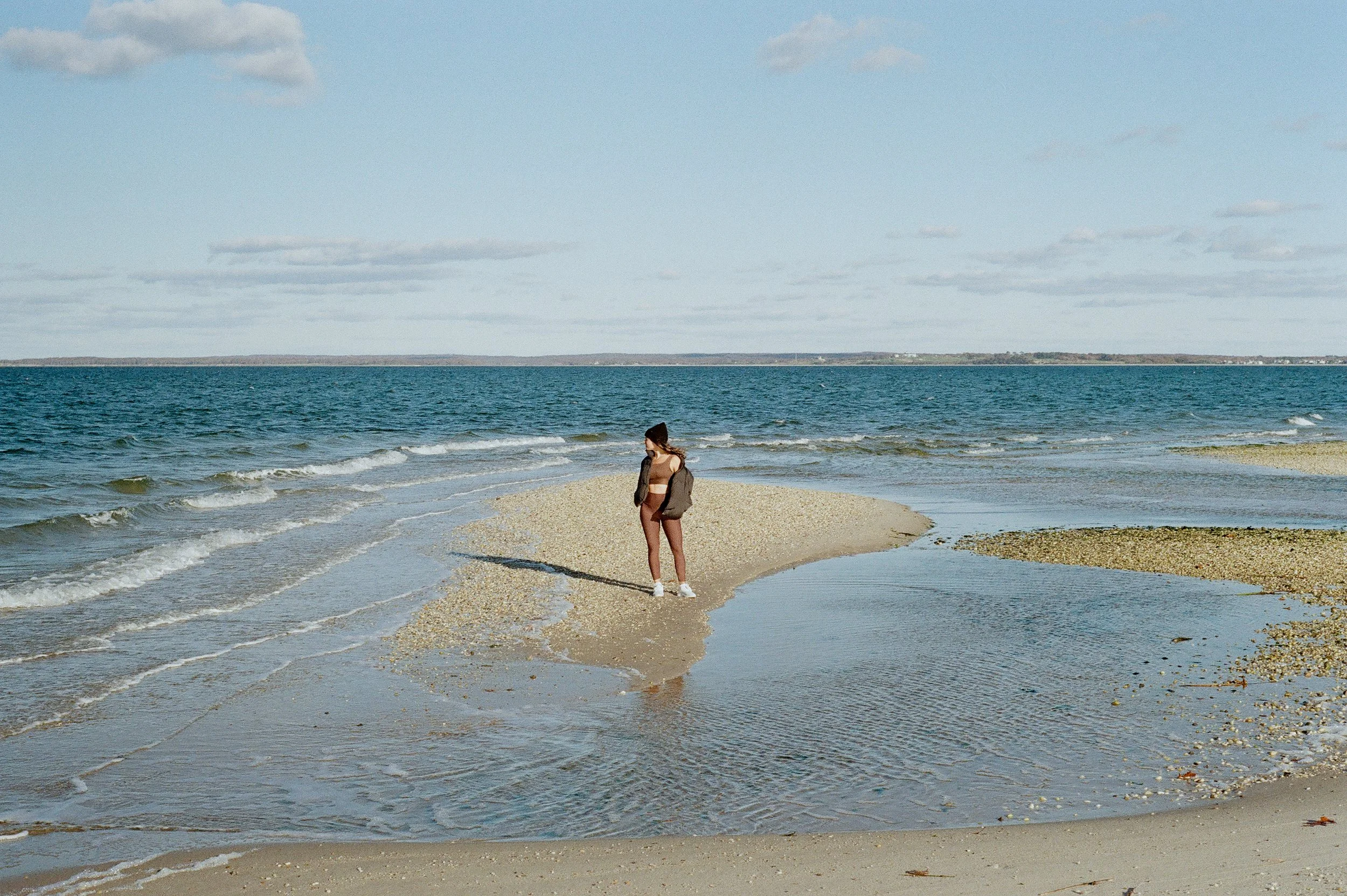 A woman standing on a sandy beach near the ocean, looking out at the water with her back to the camera. She is wearing a brown bikini, a beanie, and has a jacket slung over her shoulder. The sky is partly cloudy, and the water has small waves.